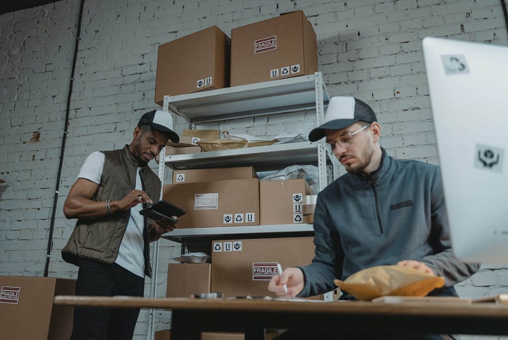 Two people in a warehouse setting, wearing caps and casual clothing. One is using a tablet near shelves stacked with cardboard boxes. The other is seated at a desk, writing on a package. A computer monitor is visible in the foreground.