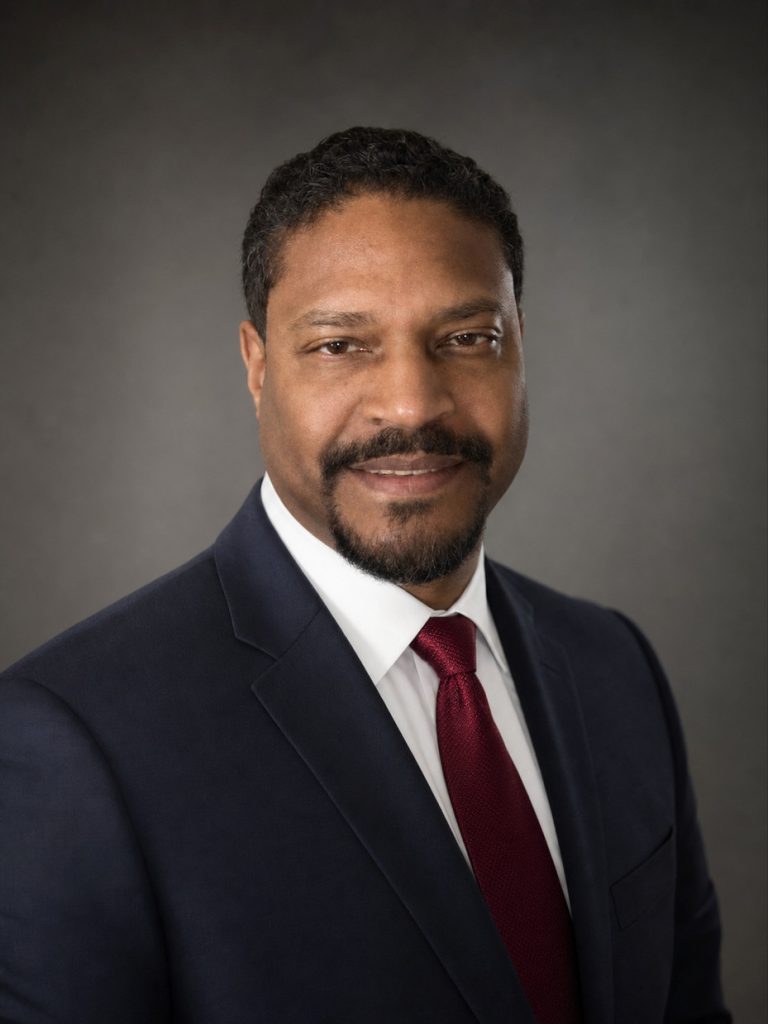 Professional headshot of a man in a navy suit, white shirt, and burgundy tie, smiling slightly against a gray backdrop.