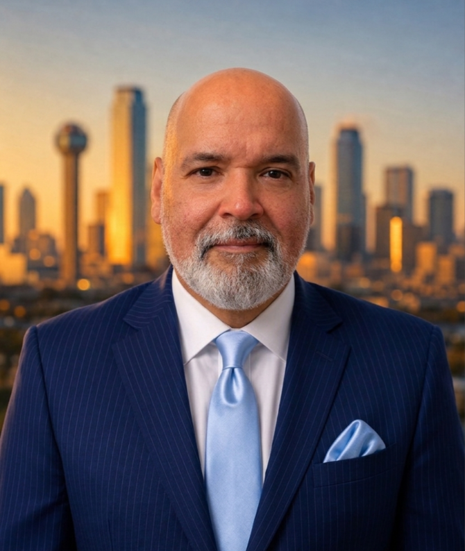 Professional head-and-shoulders portrait of a bald man with a gray beard in a navy pinstripe suit, light blue tie, and matching pocket square, with a city skyline at sunset behind him.