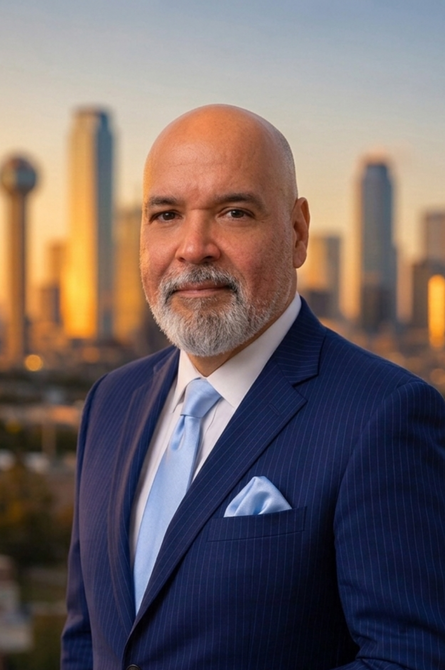 Professional head-and-shoulders portrait of a bald man with a gray beard in a navy pinstripe suit, light blue tie, and pocket square, with a blurred city skyline at sunset behind him.